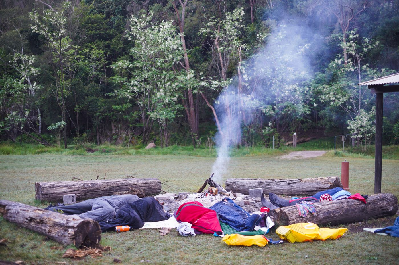 Sleeping bags and logs around a smoking campfire on a riverbank clearing