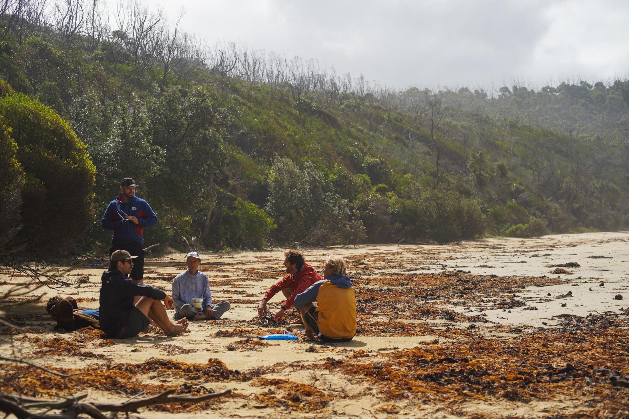 Five men sitting in a circle talking on a wild beach below misty hills