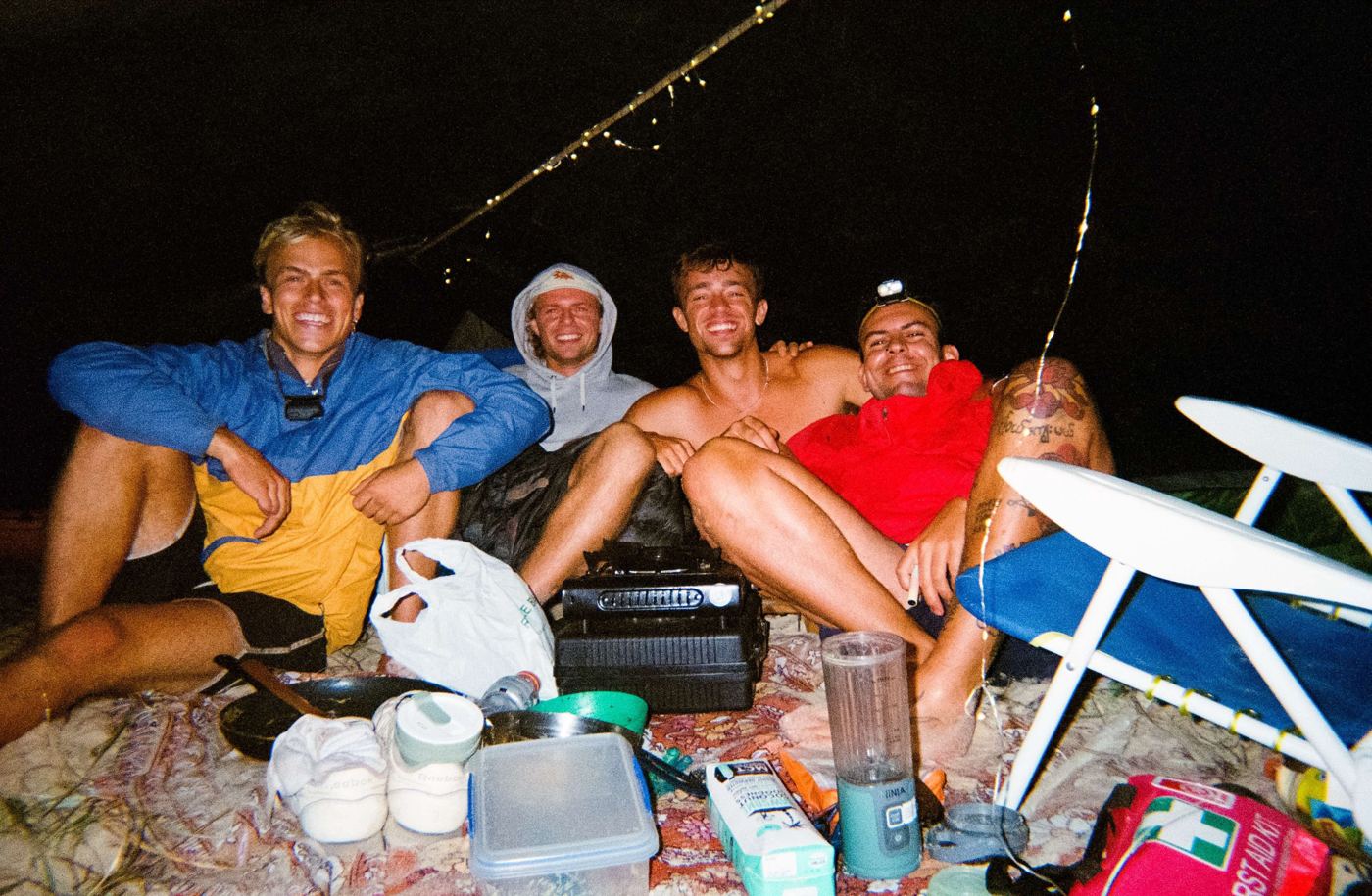 Four men laughing at camp at night under fairy lights