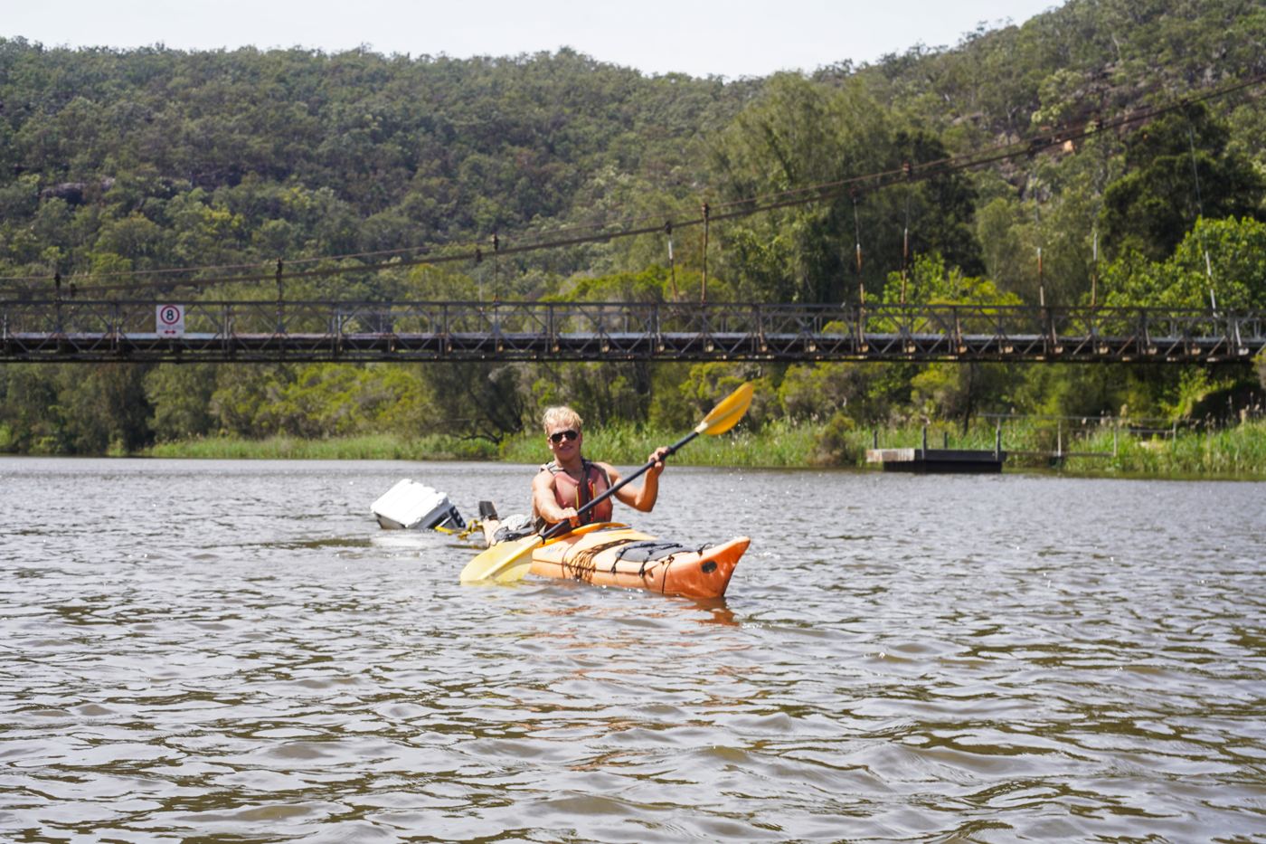 A man paddling a yellow kayak below a suspension bridge