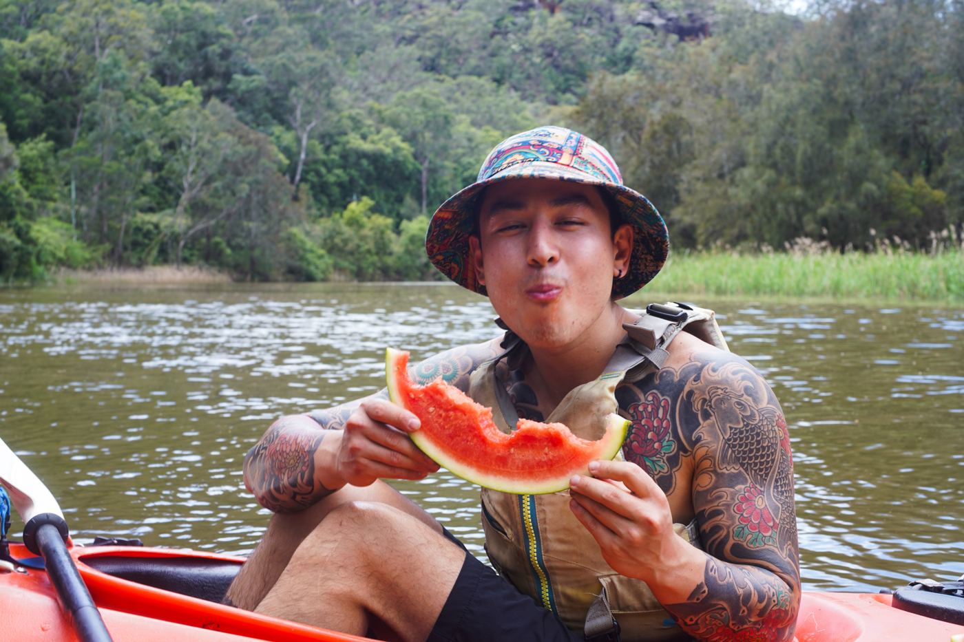 A man in a bucket hat biting into watermelon in a kayak
